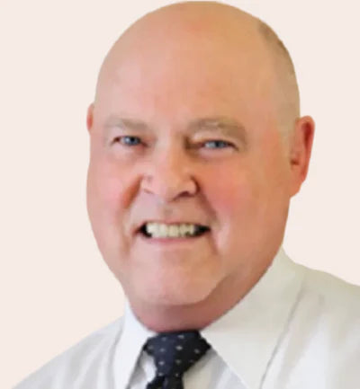 A close-up portrait of a smiling bald man with blue eyes, wearing a white collared shirt and a dark patterned tie, against a plain light background.