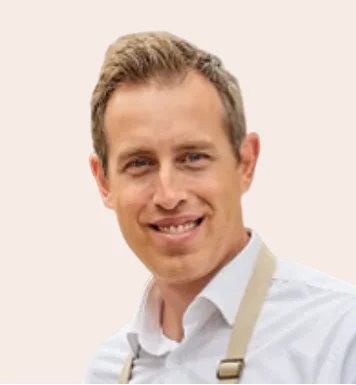 A smiling man with short light brown hair, wearing a white button-down shirt and beige suspenders, photographed against a plain light beige background.