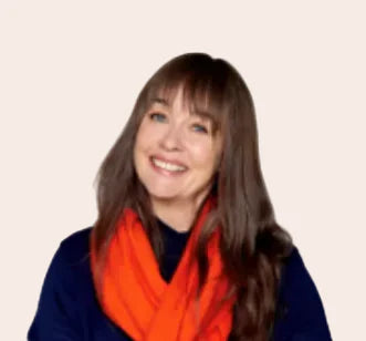 A smiling woman with long brown hair and bangs, wearing a bright orange scarf and a navy blue top, photographed against a plain light beige background.