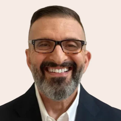 A smiling man with a graying beard and black-rimmed glasses, wearing a dark suit jacket and white collared shirt against a plain light background.